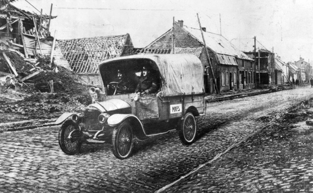 A First World War maps truck - it is more like a converted old-fashioned car, with a tarpaulin cover, being driven down a street lined by heavily damaged buildings