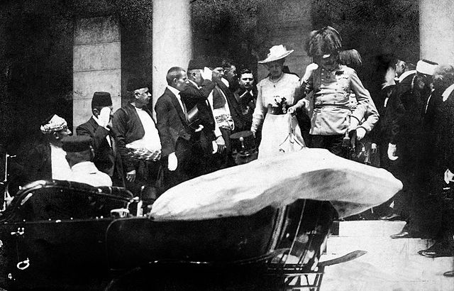 Archduke Franz Ferdinand accompanied by a women, walking down the steps to a car. He is being saluted by staff.