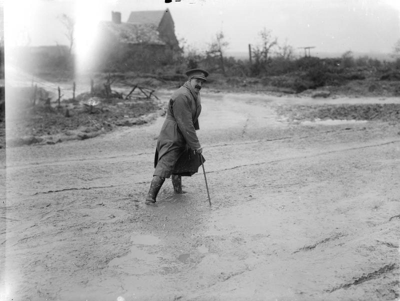 First Official War Artist Muirhead Bone crossing a muddy river, dressed in trench coat with scarf and walking cane. Maricourt, September1916.