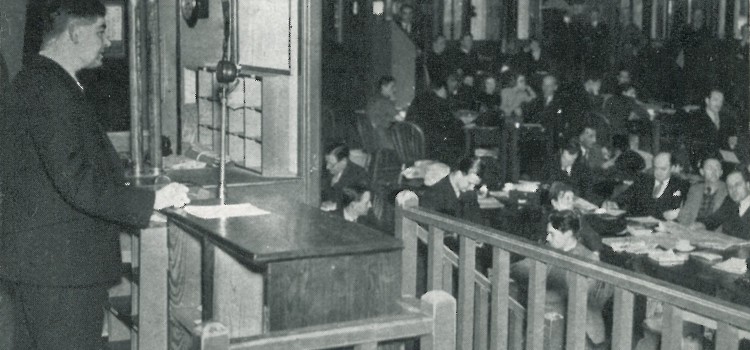 In the Ministry of Information Press Room, a man stands at a microphone, possibly making an announcement to the large number of journalists seated in the hall.