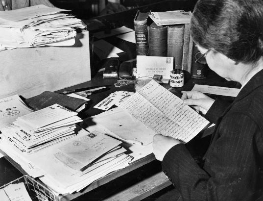 A censorship worker is inently reading a letter at a desk which bears piles of letters in envelopes to be opened