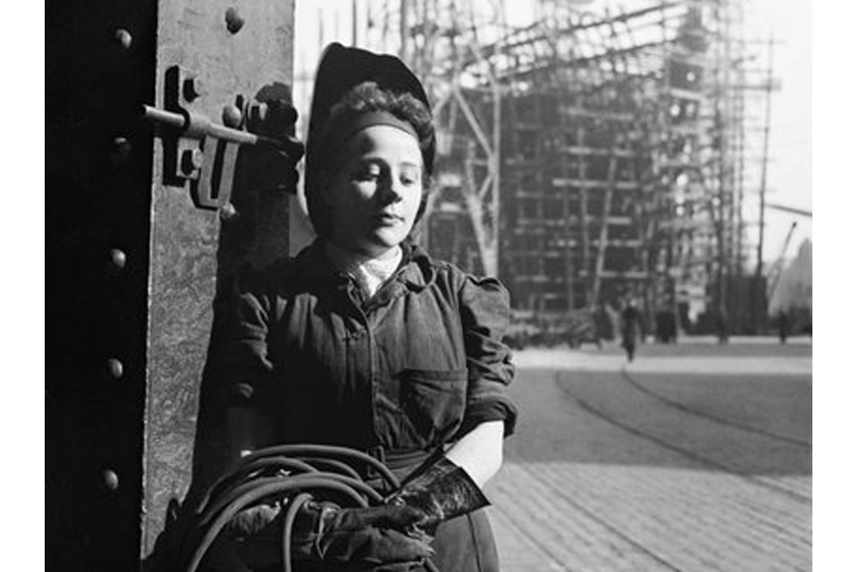 A study of a young woman welder. In the background a ship under construction in a slipway can be seen.