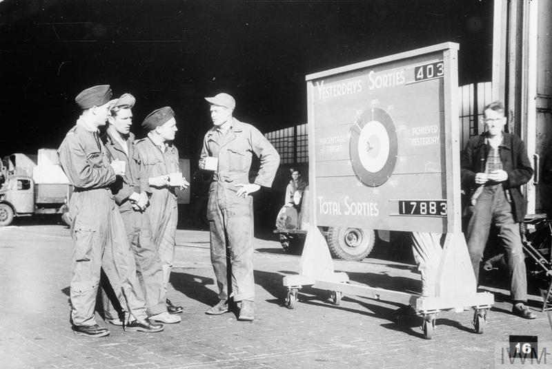 British and United States air crew take a tea break outside a maintenance hangar, probably at RAF Gatow, Berlin. Nearby, a sign displays the number of sorties carried out the day before and the overall total for the airlift.