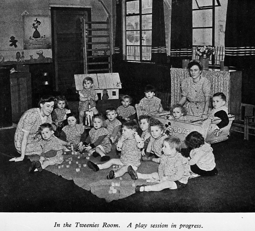 Picture is captioned: 'In the Tweenies Room. A play session is in progress'. Two female nursery staff are seated on the floor with the nursery children who are also sitting. Most of the children are on a mat, with some scattered building blocks. Three children are in a toy boat. 