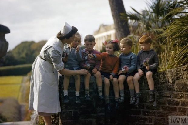 A nurse with child evacuees from Plymouth in the garden of the Chaim Weizmann Home at Tapley Park, Instow, North Devon, October 1942 Five children are shown sitting on top of a wall (two girls, three boys), and a girl in the centre is giving a posy of flowers to a nurse who is wearing a uniform.