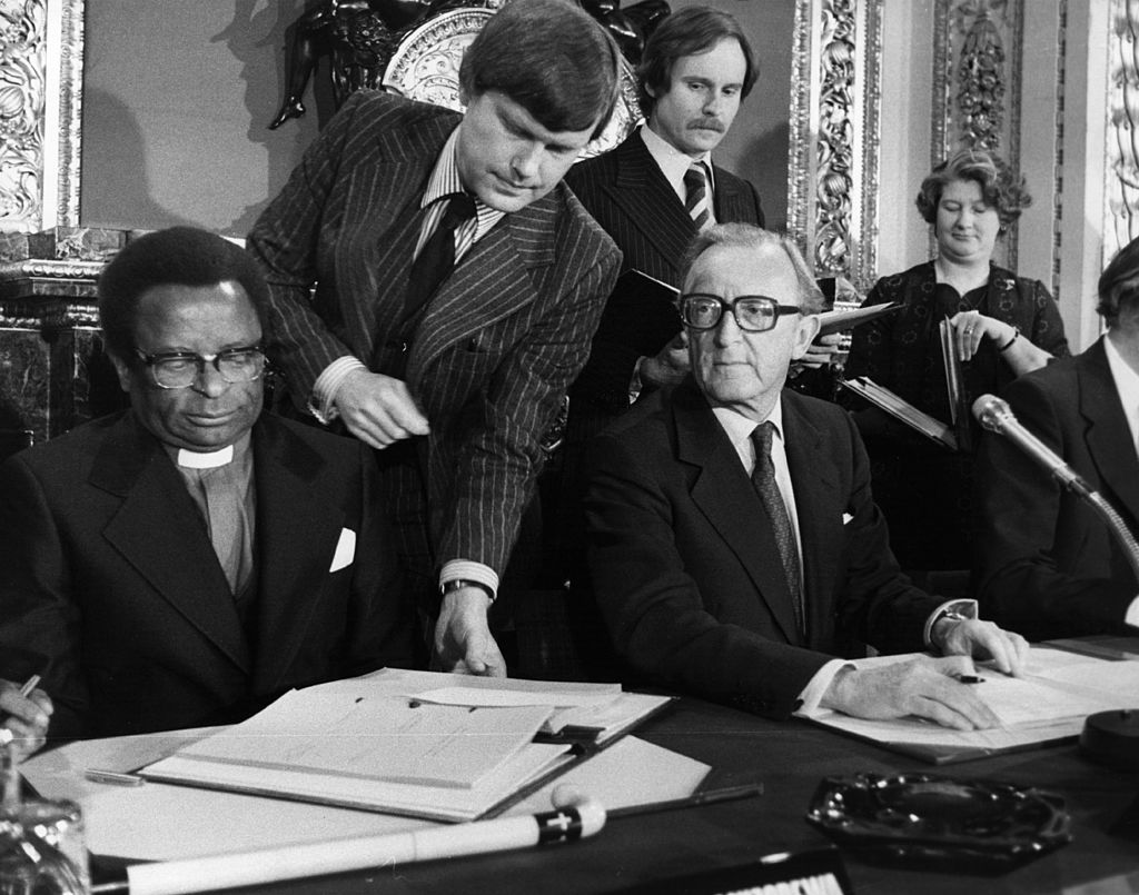 A busy scene with lots of people milling around, Methodist Bishop Abel Muzorewa and British Foreign Secretary Peter Carington are sat and are in the process of signing pact. A man is lent between them picking up the pact. 