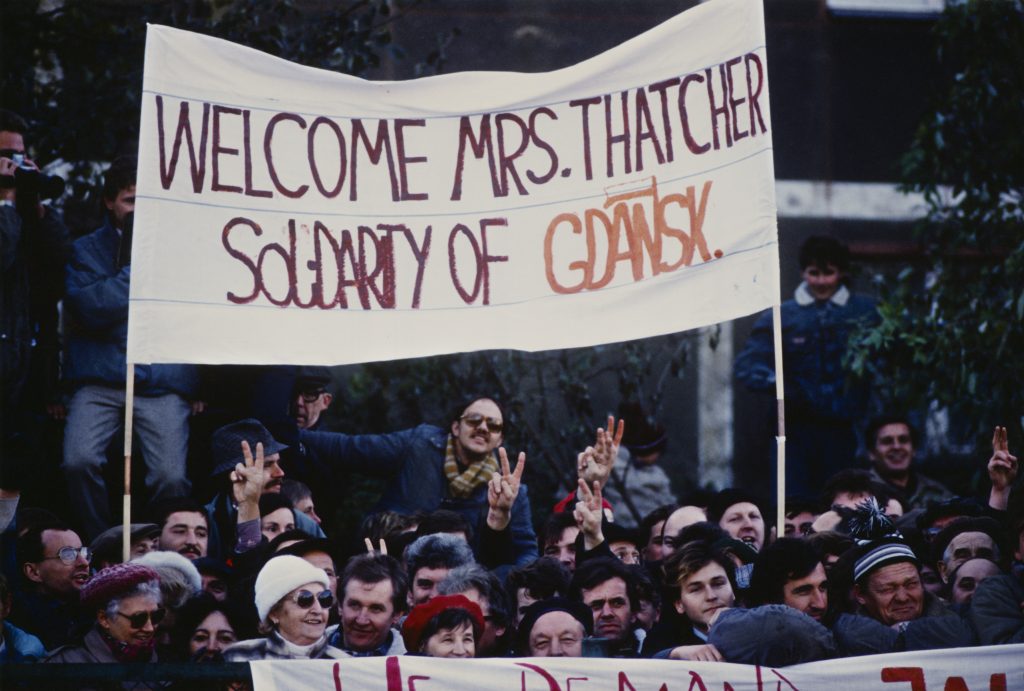 A crowd of people holding up a banner saying: Welcome Mrs. Thatcher Soldarity of Gdansk. People are also holding up peace signs.